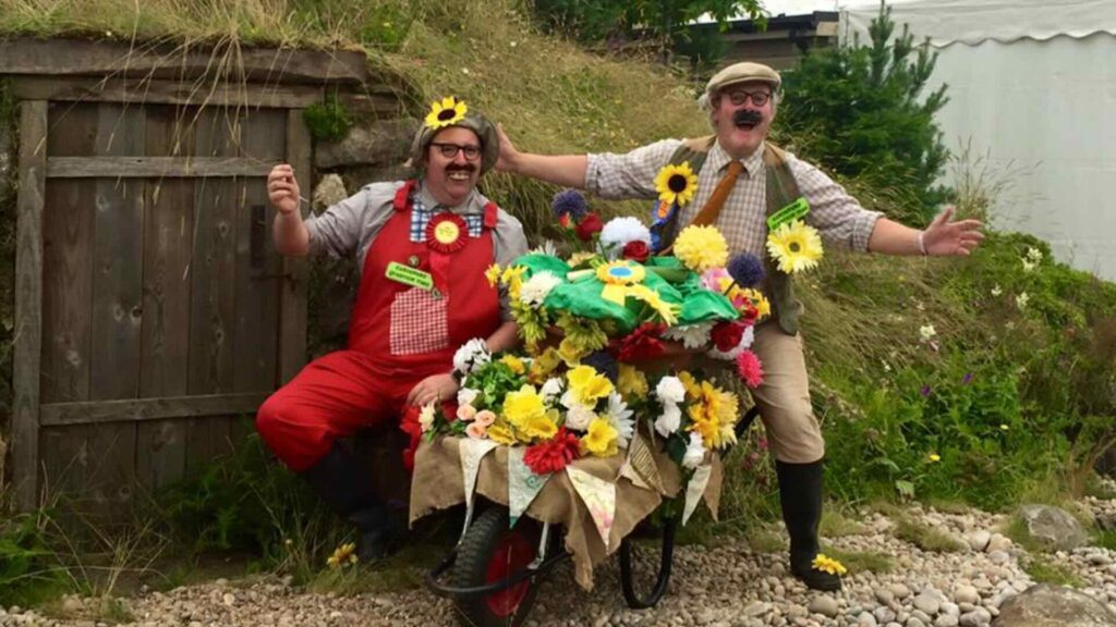 A comedy duo the avid gardeners, posing with flowers at an outdoor event.