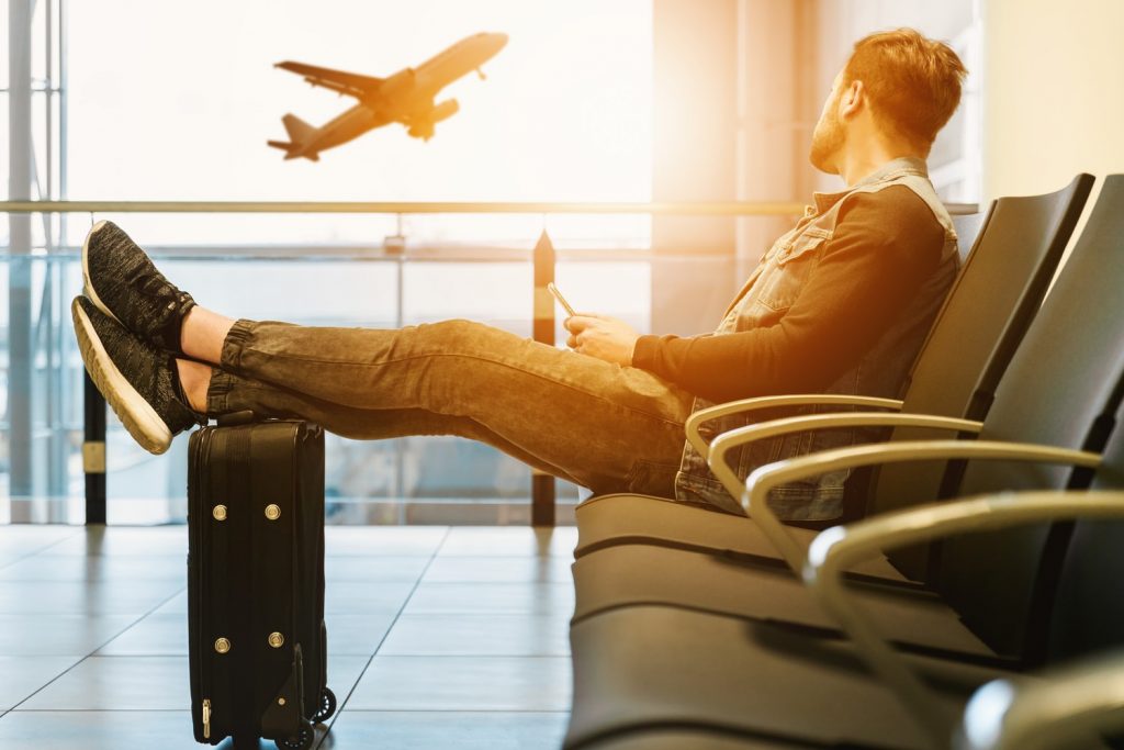 Young man from an Events Agency sitting on an airport seat in an airport terminal watching a plane tack off in front of the sun.