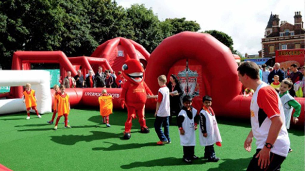 Children playing in Liverpool FC inflatable football pitch with dragon mascot hired as part of football themed family fun day