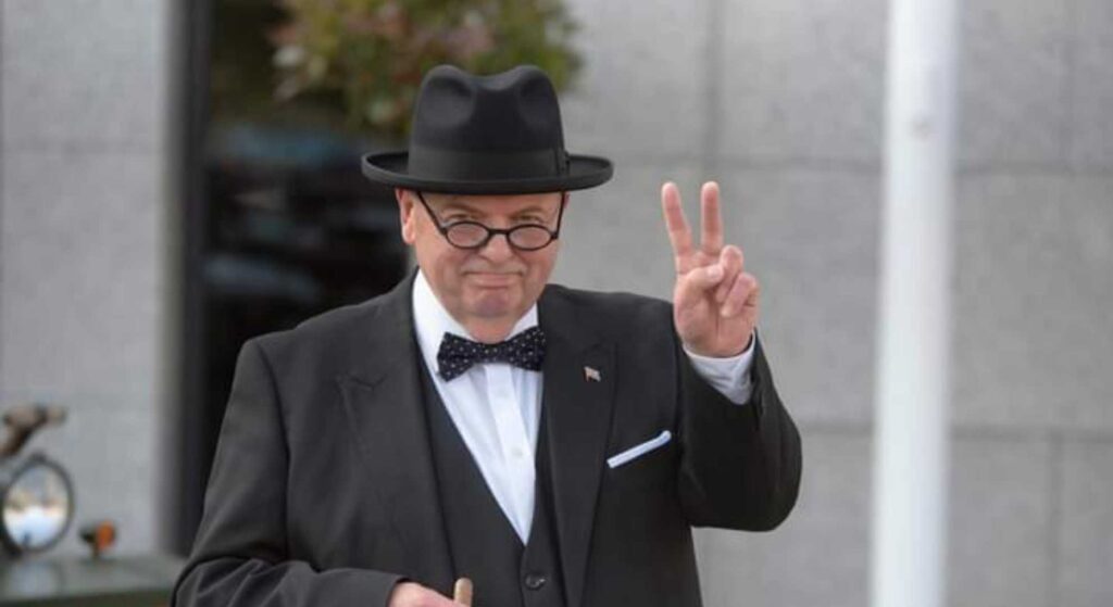 A Winston Churchill impersonator performing at a live event in smart black attire, with two fingers up in the Churchill famous gesture.
