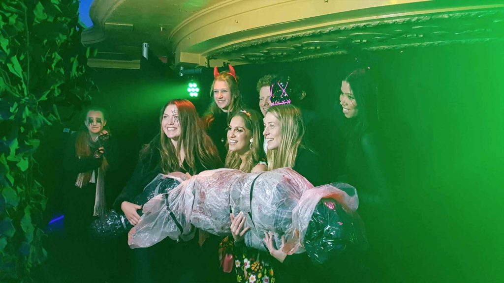Group of friends at Halloween themed event posing in photo booth area holding giant Halloween body prop.