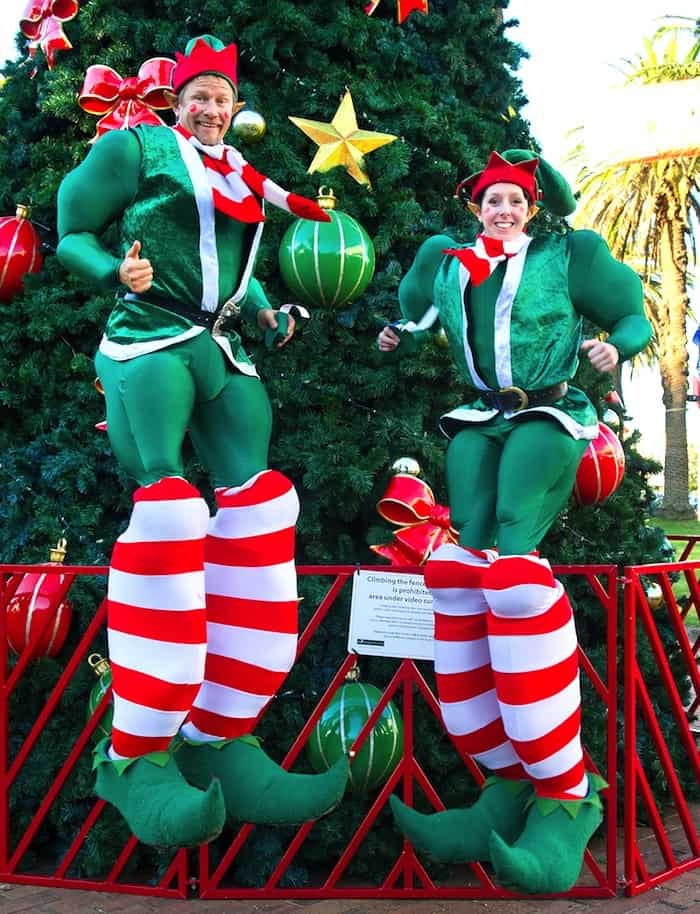 Bouncing Elf Stilt Walkers standing in front of a large Christmas Tree at a Santa Grotto event in a Garden Centre in the UK.