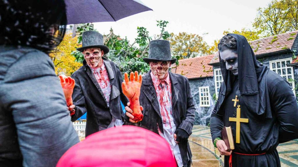 Two scare actors in hats and priest scaring audiences with hand props in scare attraction event.