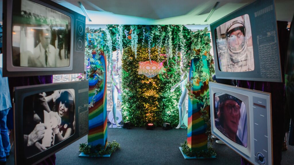 Colourful archway with fake foliage and retro TV props either side; corporate set design to 1960s themed event.