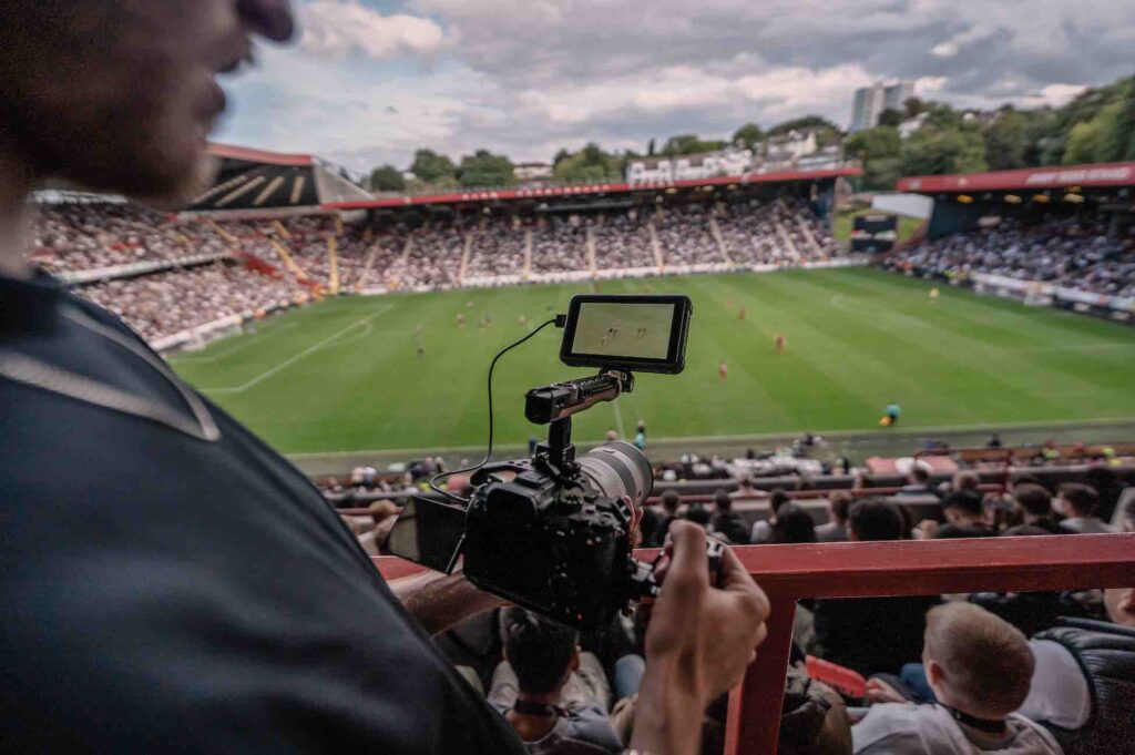 Videographer capturing footage at football match in stands.