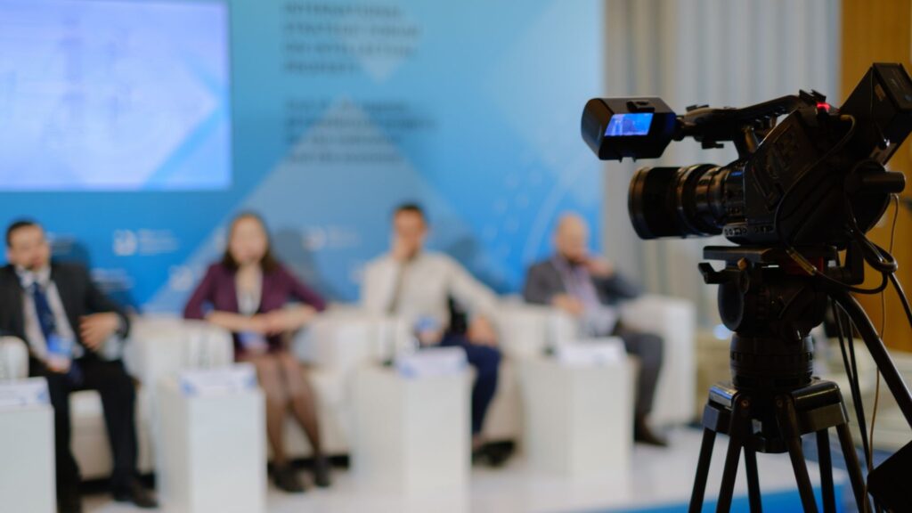 Camera on tripod recording guest speakers at conference.