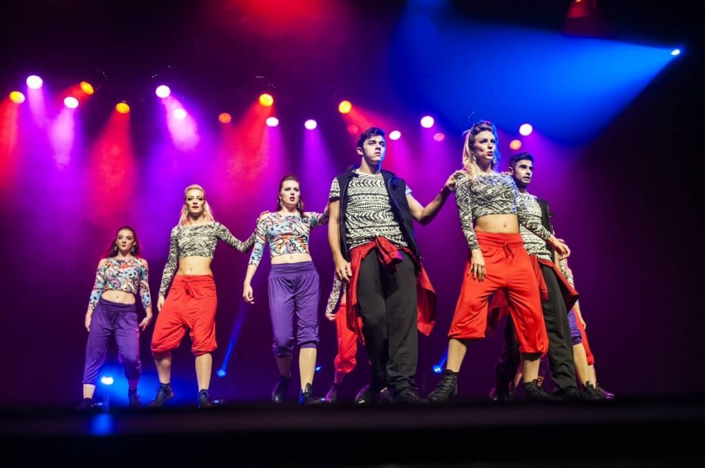 Male and female breakdancers standing in a v shaped unit on performing on stage at festival themed party wearing street fashion clothing.