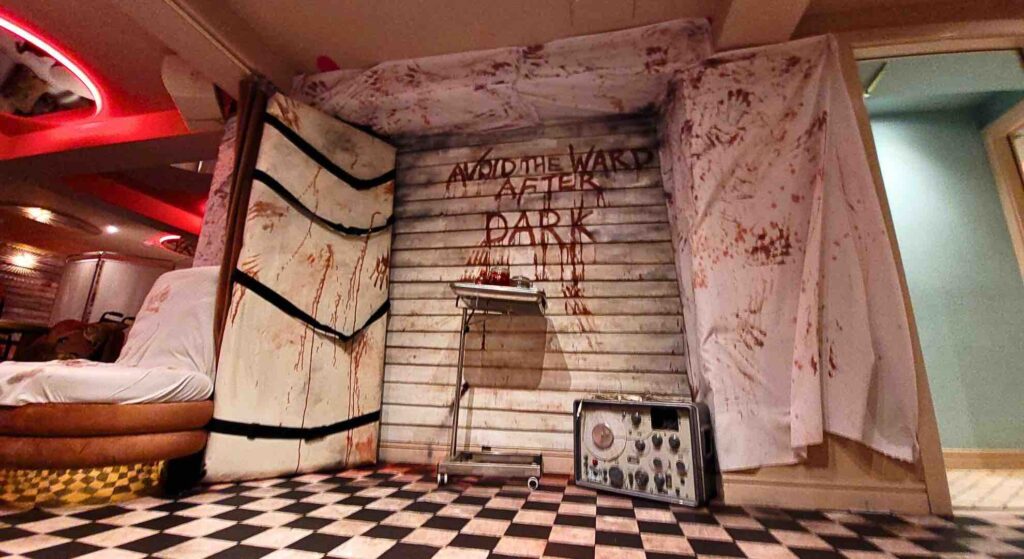 The "AVOID THE WARD AFTER DARK" set design features a gory medical tray, ominous table with straps, and a chilling electrolysis machine, setting a nightmarish tone.