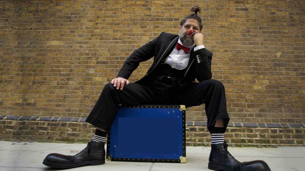 Clown sits on suit case with bored expression