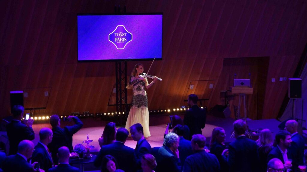 Crowd watches a violinist performing on stage, lit in a purple light and wearing a white and black dress.