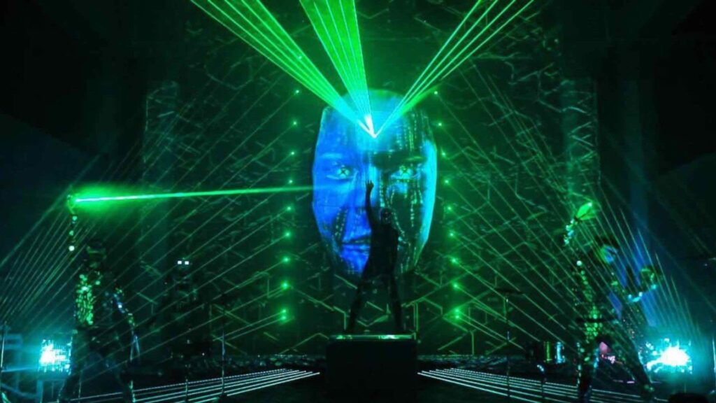 Projection mapped face with green laser show being projected onto drummers performing on stage