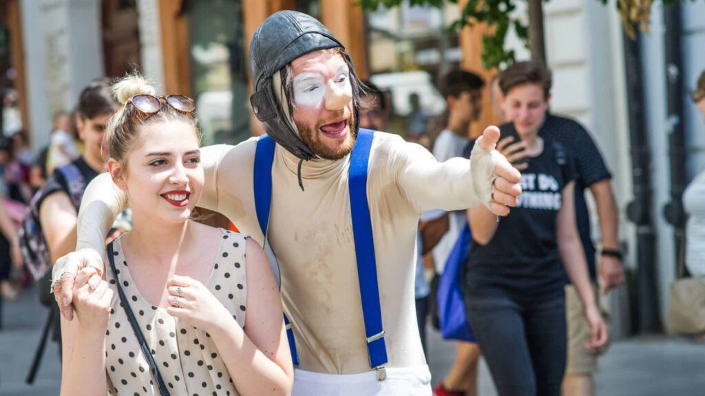 A street theatre performer wraps his arm around a woman whilst gesturing to his counterpart.