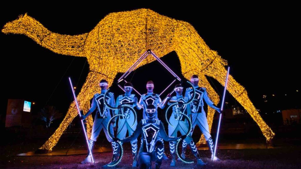 A troupe of LED poi dancers stand on stage in LED costumed with a camel prop