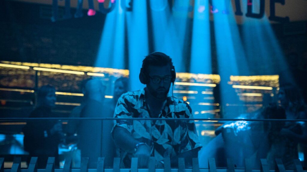 A DJ mixes music in a club with blue beams on him