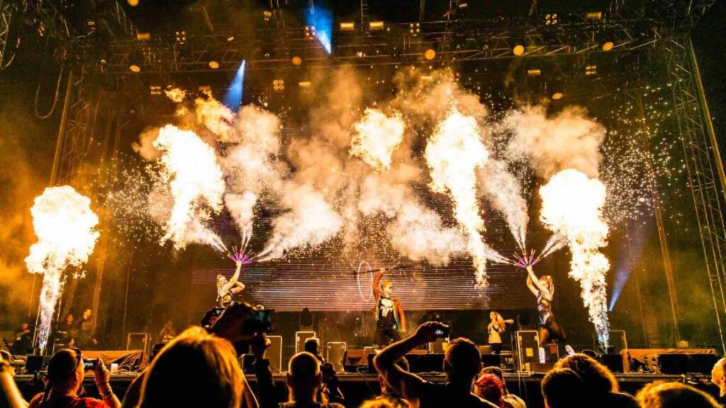 Performers holding fire fans with large flames bursting into the air while performing on stage with flames bursting around them.