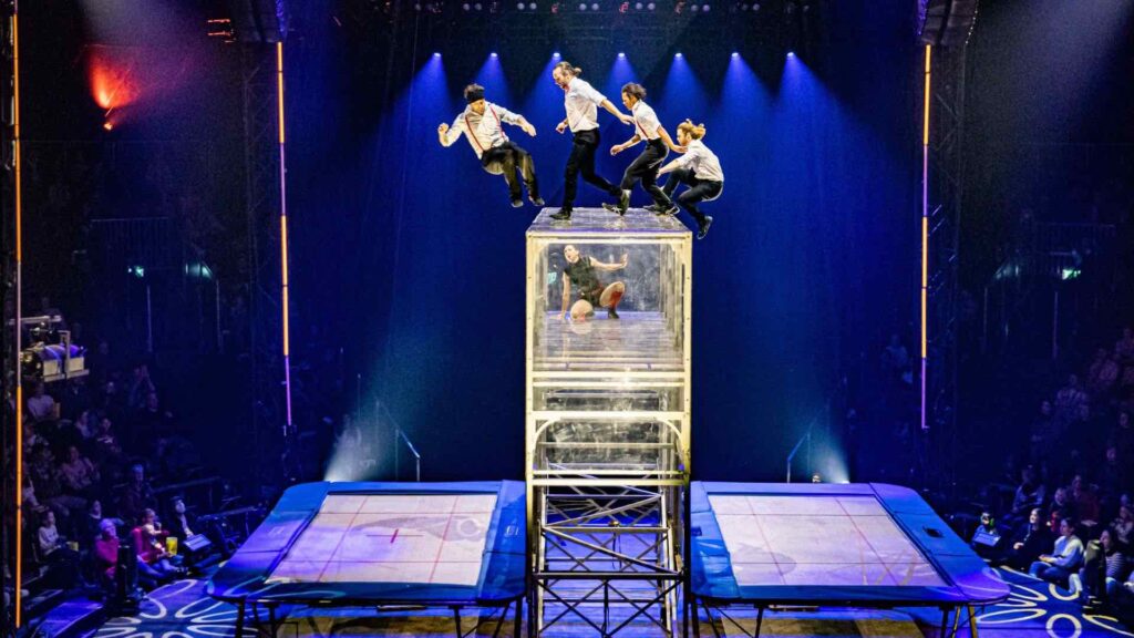 Four acrobats jump over a tall transparent wall using trampolines