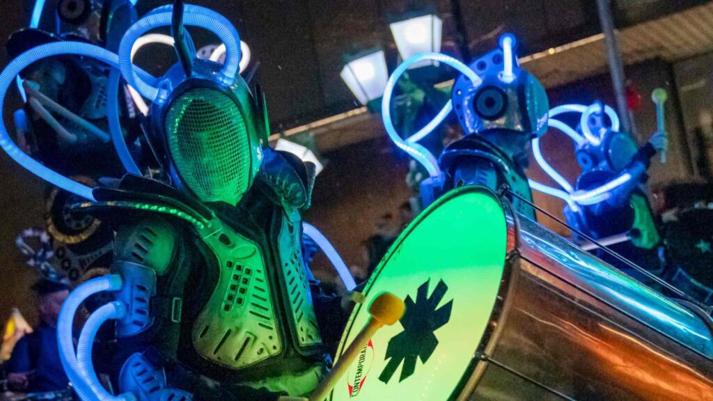 A glow bot in a green and blue costume is drumming with LEDS