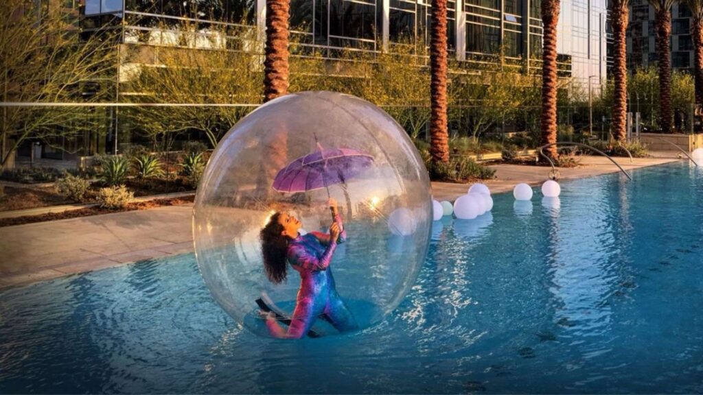 A lady inside a giant bubble dome floating on water performing acrobats with an umbrella with a palm tree backdrop