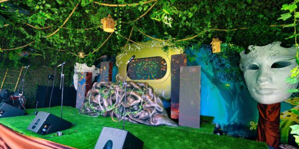 Whimsical forest-themed stage with green canopy, hanging birdhouses, microphones, speakers and a large white mask.