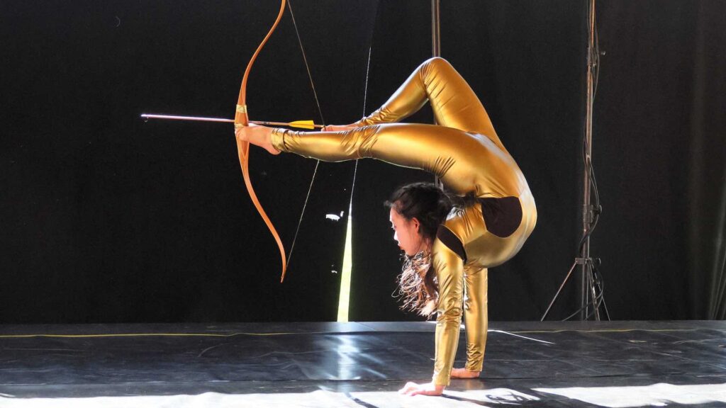 A female contortionist bends with a bow and arrow in her feet ready to hit a target
