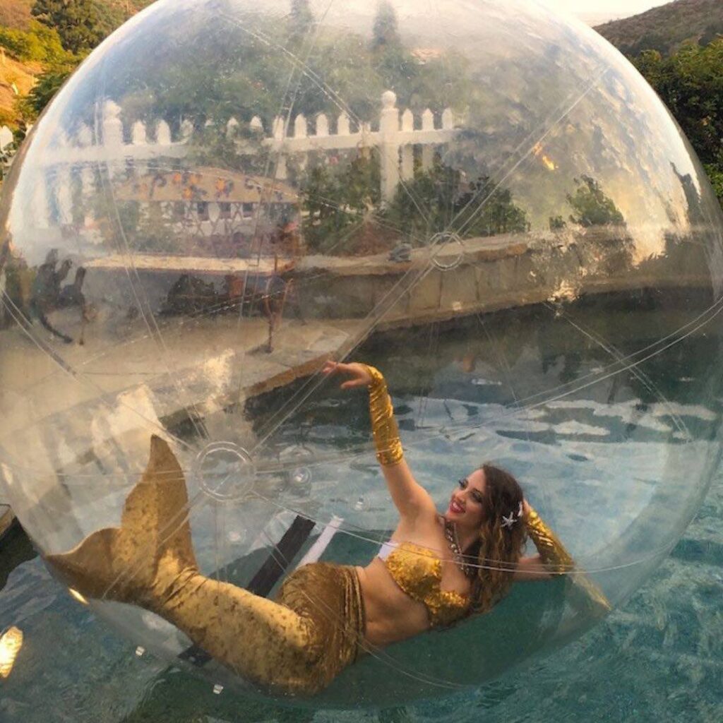 A lady in a mermaid costume inside a giant bubble sphere which sits on water where she acts as a focal point