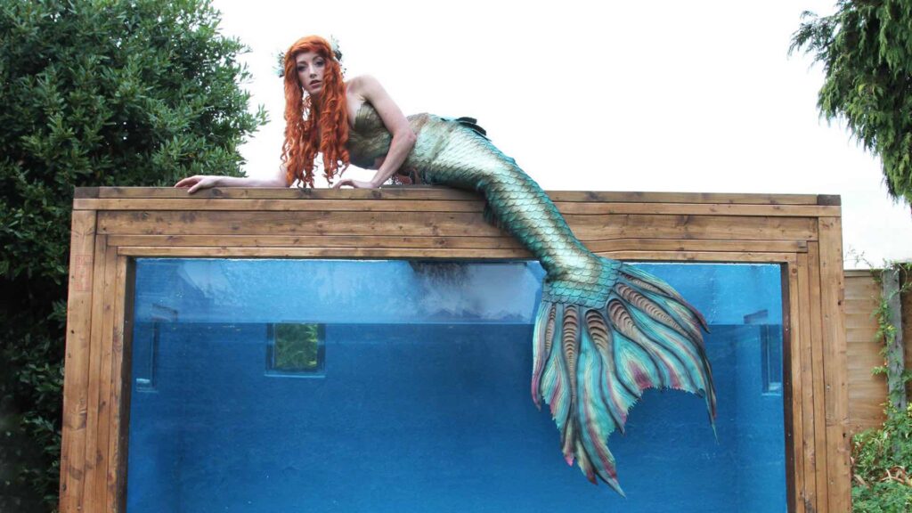A mermaid lays on top of a mermaid tank bespoke built for mermaid performances