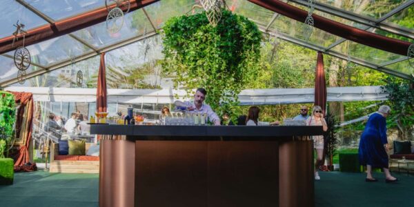 Bartender serving drinks at a round bar under a clear tent with suspended greenery.
