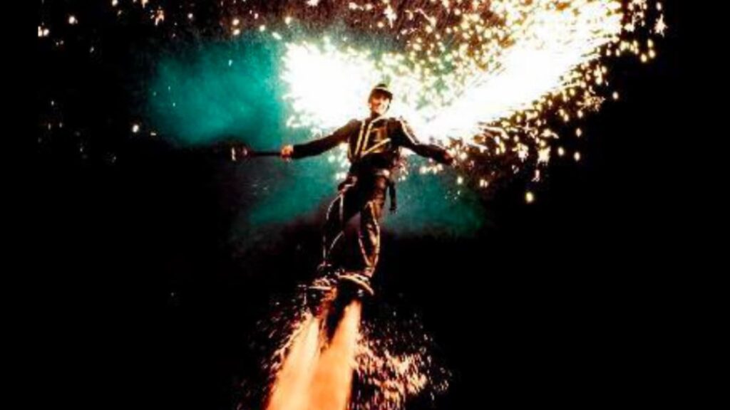 Flyboard performer airborne above the water with fireworks sparking from the board.