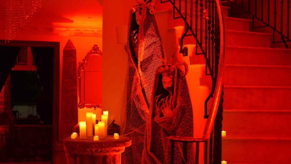 Bespoke mysterious statue props in hooded capes, holding candles at the bottom of spiral staircase in hallway area of Halloween mansion party.