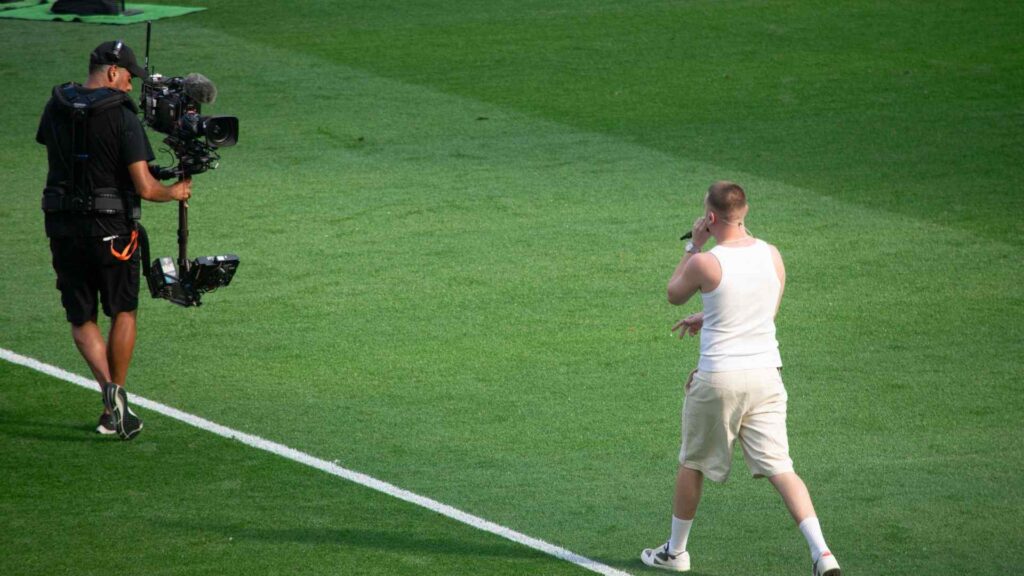 Aitch walking on pitch to centre circle for half time performance, being filmed by camera man in front.
