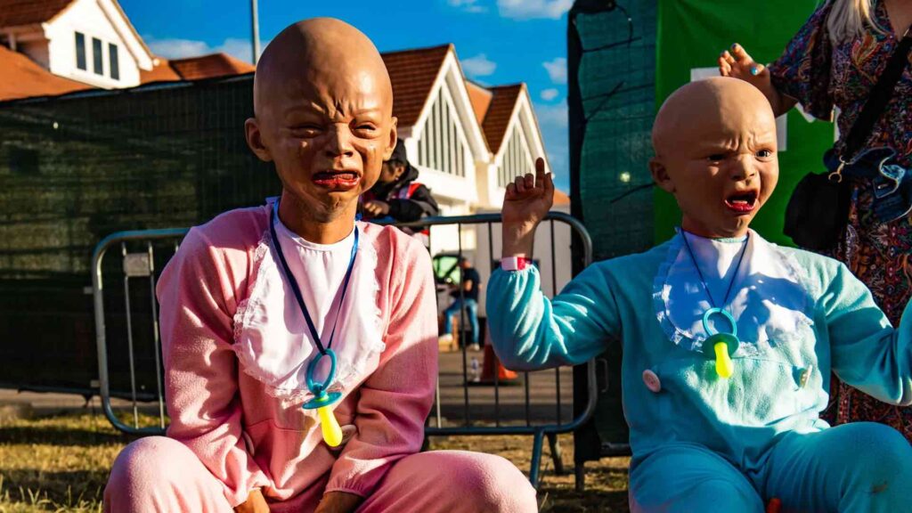 Duo of models wearing crying baby head masks in onesies at an outdoor event.