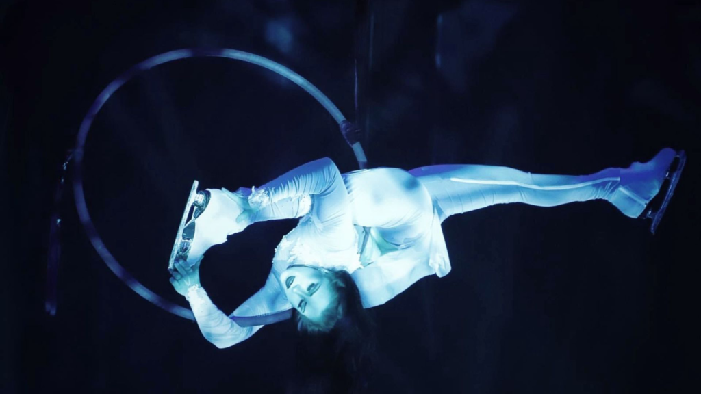 Aerial pyrotechnics ice skater hanging from hoop in the air while wearing ice skates.