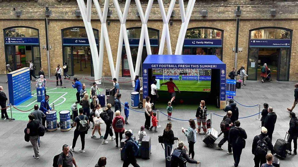 Crowd gathered at Kings Cross train station watching guests play football simulator.