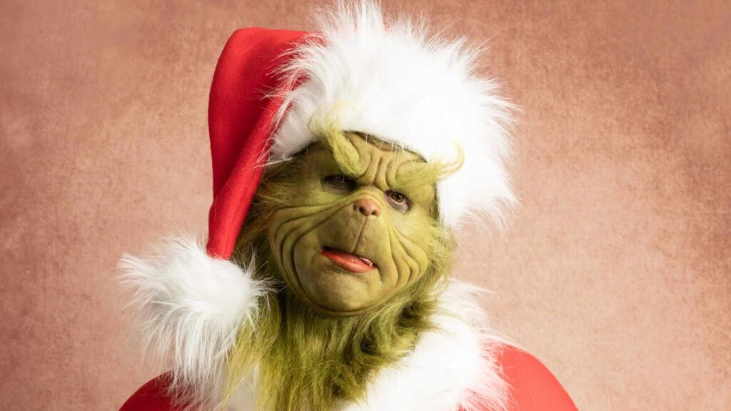 A realistic person dressed as The Grinch, posing with a red fluffy Christmas hat and looking annoyed