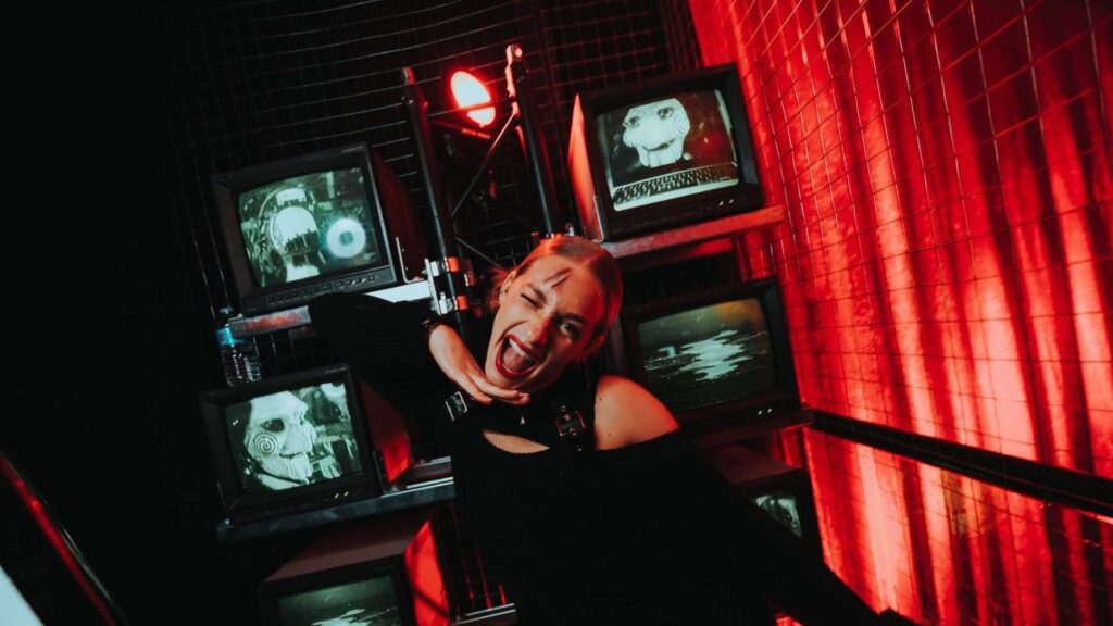Girl posing with silly face infront of television stack with creepy billy the puppet on at a SAW movie event.