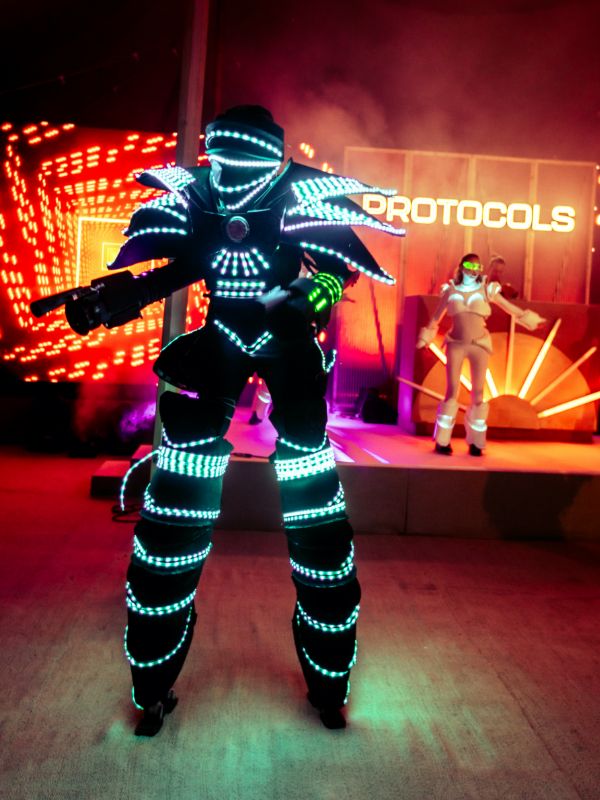 Green glow bot on stage with glow girl in the background at corporate festival.