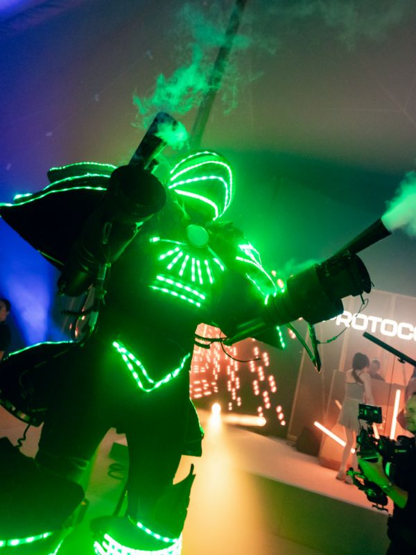 Green glow bot led dancer with smoke machine barrels as arms at corporate festival.