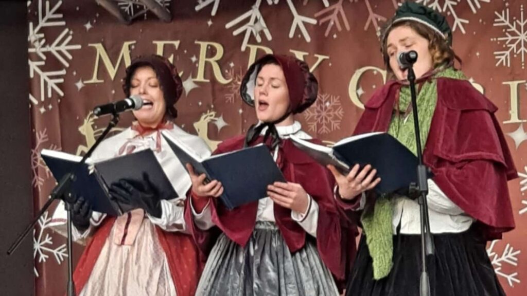 Trio of traditional Christmas carol singers in Victorian style outfits performing at a public Christmas event.