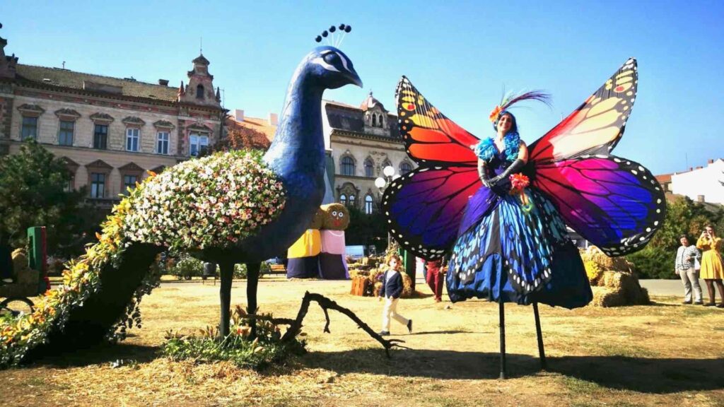 A giant butterfly stilt walker stood next to a large bush peacock