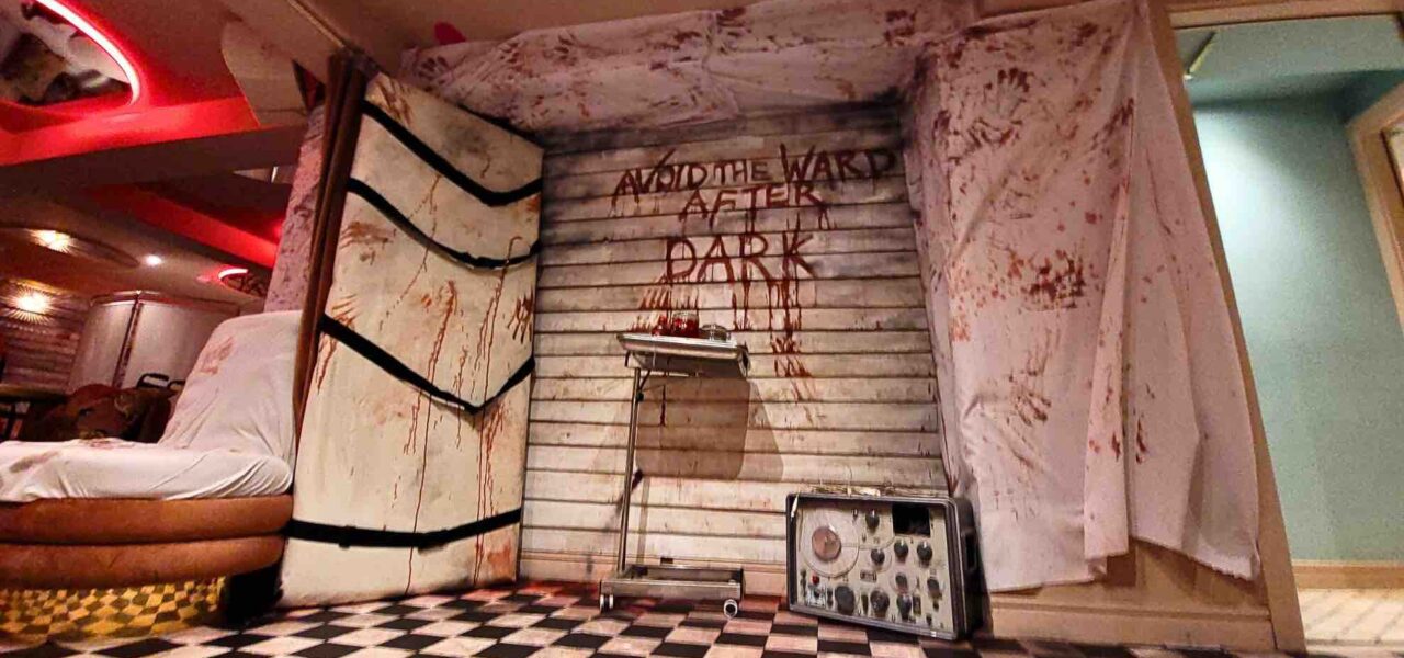 The "AVOID THE WARD AFTER DARK" set design features a gory medical tray, ominous table with straps, and a chilling electrolysis machine, setting a nightmarish tone.
