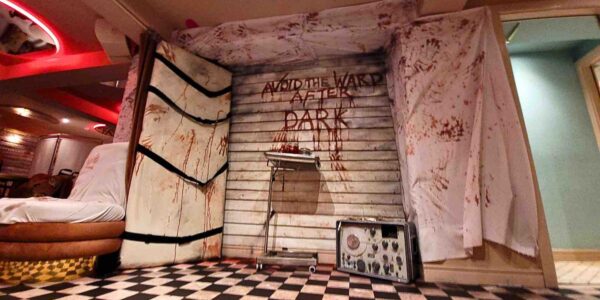 The "AVOID THE WARD AFTER DARK" set design features a gory medical tray, ominous table with straps, and a chilling electrolysis machine, setting a nightmarish tone.