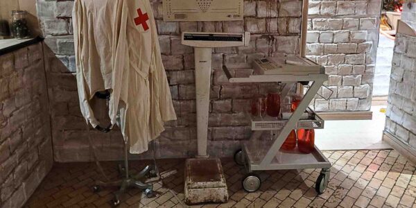 A disturbing medical setup featuring an old exam table, weighing scale, hanging aprons, and an optician’s sign evokes a sinister clinical scene.