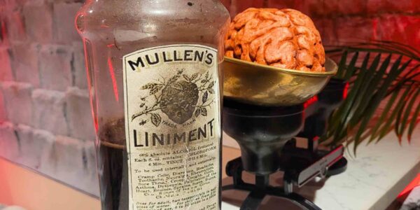 A jar of "Mullen’s Liniment" sits beside a brain medicine prop on an old scale, creating an unsettling scene of medical horror in the Asylum-themed setup.
