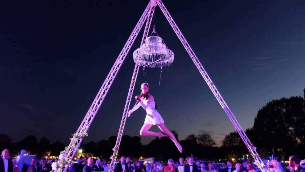 An aerial violinist hanging from a chandelier at an outdoor event with rigging.