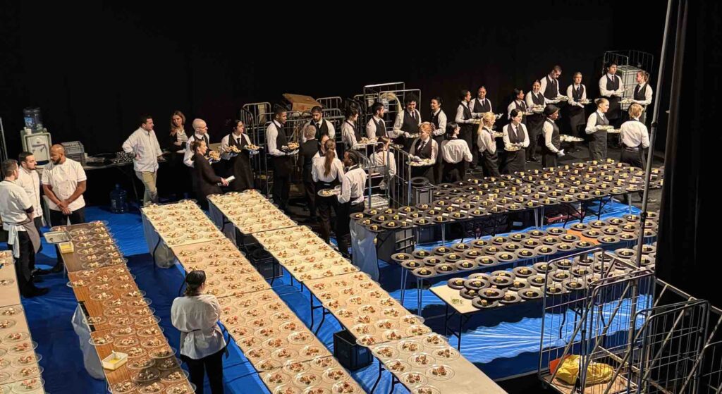 Behind the scenes of mass catering at a gala event in London. Showing lots of plates of foods laid on tables with catering staff being delegated orders.