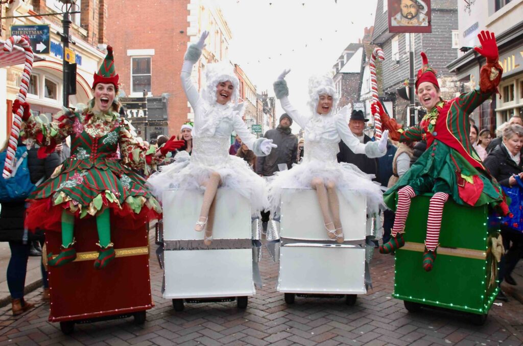 Festive Christmas angels and elfs sitting on fake presents form a group of LED performers at a festive parade.