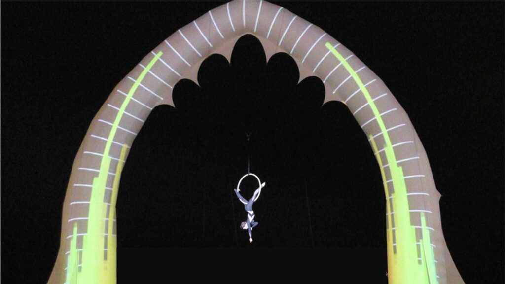 An aerialist hanging from a hoop attached to an LED arch performing at an evening event for Christmas.
