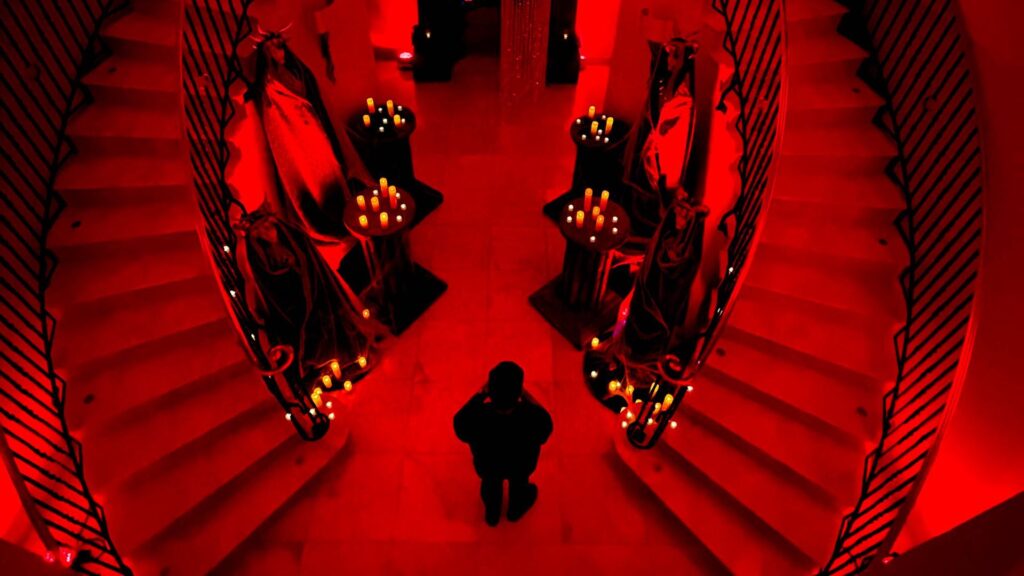 Two spiralling staircases lit in red lighting, with candles and a dark figure.