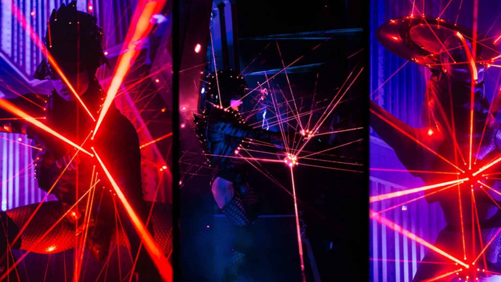 Pair of laser performers on a dark stage surrounded by smoke and coloured beams.