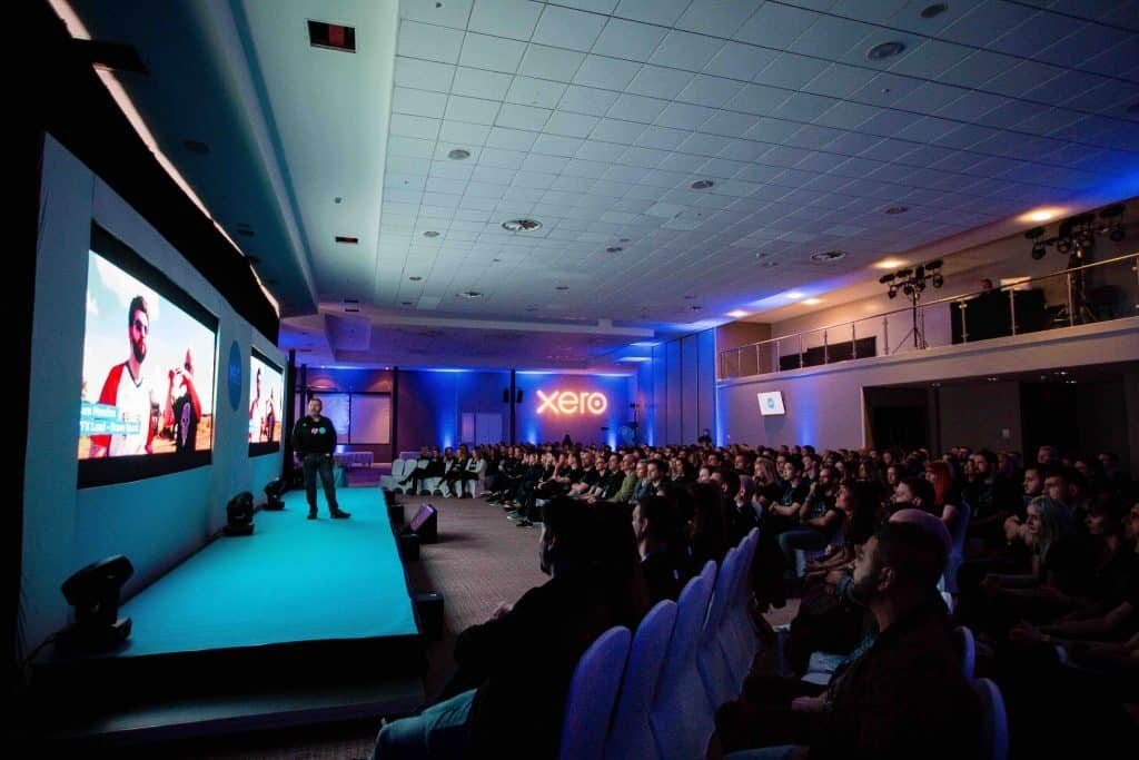 A dark room illuminated by a light-up Xero sign, LED screens behind a stage and a speaker talking to the crowd.